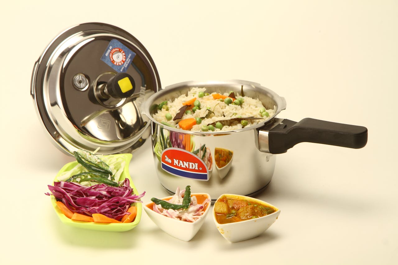 Cooked Indian dish in a Nandi pressure cooker with side dishes on white background.